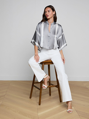 A woman with long brown hair sits in a black chair, wearing the Dru Silk Blouse with kimono sleeves, paired with white pants and high-heeled sandals. The setting has a light wall and wooden floor.