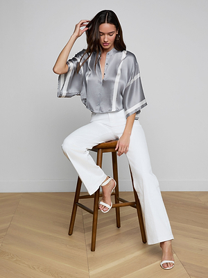 A woman with long brown hair sits on a black armchair, wearing the Dru Silk Blouse, paired with white pants and white heeled sandals. She faces the camera with a neutral expression against a plain background.
