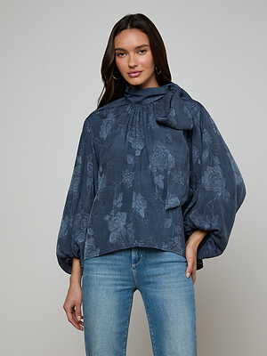 A woman with long dark hair wears the Rhett Oversized Blouse, featuring blue floral jacquard, voluminous sleeves, and a bow at the neck, paired with light blue jeans against a plain light background.