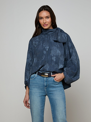 A woman with long dark hair wears the Rhett Oversized Blouse, featuring a floral jacquard design, voluminous sleeves, and neck tie, paired with light blue jeans and a black belt against a plain light background.