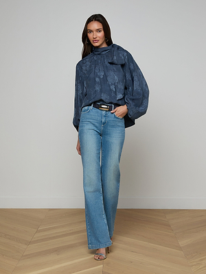 A woman with long brown hair stands indoors on a wooden floor, wearing the Rhett Oversized Blouse with a neck bow, paired with light blue wide-leg jeans, a black belt, and heeled sandals. She appears relaxed.