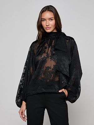 A woman with long brown hair wears the L'AGENCE Rhett Burnout Satin Blouse, a sheer black floral top featuring a large bow at the neck and billowy sleeves, paired with black pants against a light gray background.