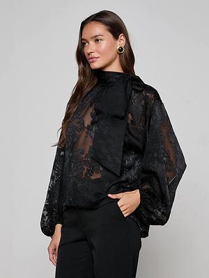 A woman with long brown hair models the L’AGENCE Rhett Burnout Satin Blouse—an oversized, sheer black floral top with a large neck bow—paired with black pants. She stands against a plain light background, facing slightly to the side.