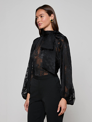 A woman poses against a gray background in the L'AGENCE Rhett Burnout Satin Blouse—an oversized sheer black floral top with voluminous sleeves and a large neck bow—paired with black pants, gold earrings, and straight brown hair.