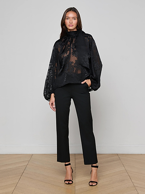 A woman poses indoors against a light-gray wall wearing the L'AGENCE Rhett Burnout Satin Blouse, featuring a sheer black finish and statement bow, styled with black straight-leg pants and high-heeled sandals. She has long brown hair and a neutral expression.
