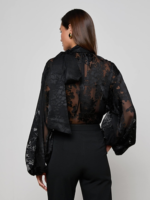 A woman with long brown hair, seen from the back, wears the L'AGENCE Rhett Burnout Satin Blouse featuring sheer black floral fabric, voluminous sleeves, and a tied neck detail, paired with high-waisted black pants.