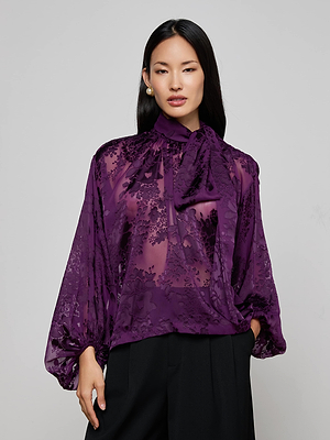 A woman wears the Rhett Burnout Satin Blouse by L'AGENCE, a sheer bohemian top with a purple floral pattern, necktie, and billowy sleeves, paired with black pants against a plain light gray background.