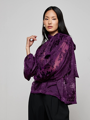 A woman with long dark hair confidently poses against a light gray background, wearing the L'AGENCE Rhett Burnout Satin Blouse in sheer purple floral with a neck bow, paired with black pants.