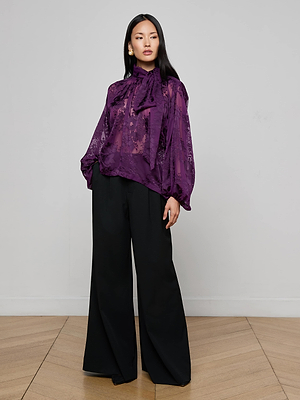 A woman poses indoors against a light gray wall, wearing the L'AGENCE Rhett Burnout Satin Blouse—a sheer purple top with high neck and bow—paired with wide-leg black pants. She has long straight hair and stands on a wooden floor.