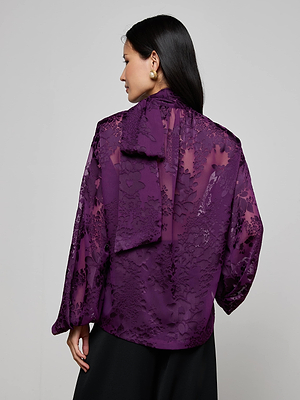 A woman with long dark hair is shown from the back wearing the Rhett Burnout Satin Blouse by L'AGENCE, a sheer purple floral top with a large tied neck bow, paired with black pants and gold earrings.
