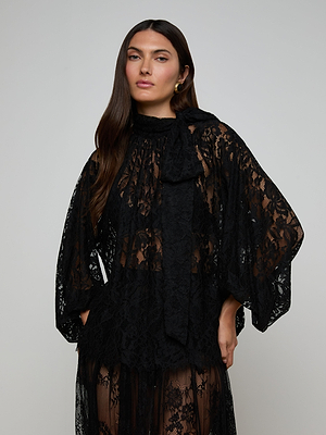 A woman with long dark hair wears the Rhett Oversized Lace Blouse, featuring sheer black floral lace, wide sleeves, and a neck bow, standing against a plain light gray background.