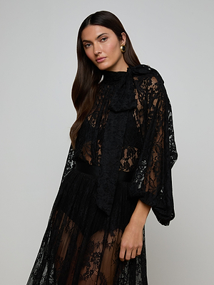 A woman with long brown hair models the Rhett Oversized Lace Blouse, featuring sheer black floral lace, a high neck with a large bow, and wide sleeves, standing before a plain light gray background.