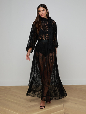 A woman with long brown hair wears the Rhett Oversized Lace Blouse, a sheer black floral lace style with long sleeves, high neck, and bow detail. She stands on a light wood floor against a white wall, wearing black high-heeled sandals.