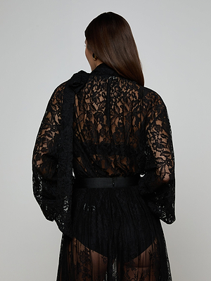 A person with long brown hair faces away, wearing the Rhett Oversized Lace Blouse as a robe with a belt and black undergarments, set against a plain light background.