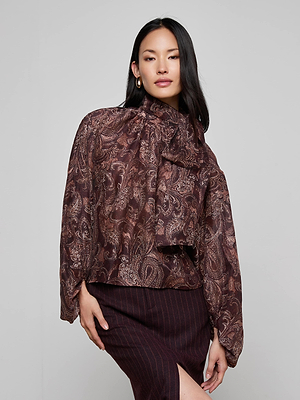 A woman models the Rhett Oversized Blouse by L'AGENCE, featuring long sleeves and a bow at the neck in a paisley print, paired with a dark thin-striped skirt against a plain light background.
