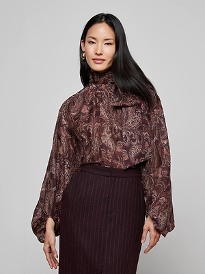 A woman with long dark hair wears the L'AGENCE Rhett Oversized Blouse, a silky bohemian paisley top with voluminous sleeves and a high neck, paired with a high-waisted dark pinstriped skirt against a light gray background.