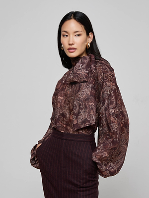 A woman with long dark hair models the Rhett Oversized Blouse by L'AGENCE, featuring a maroon paisley print, voluminous sleeves, and a bow neck. She pairs it with a high-waisted pinstripe skirt and looks to the side against a plain background.