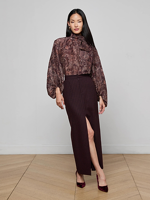 A woman indoors wears the L'AGENCE Rhett Oversized Blouse with a sheer paisley print, voluminous sleeves, and high neck, paired with a dark high-waisted skirt with front slit and maroon heels.
