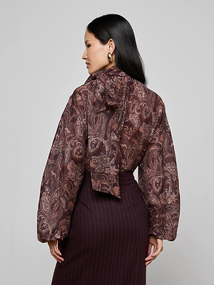 A woman with long dark hair and gold hoop earrings is shown from behind in the L'AGENCE Rhett Oversized Blouse, a brown paisley print top with a back bow, paired with a dark skirt against a plain light background.