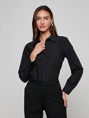 A woman with long dark hair stands against a plain background, wearing L'AGENCE's Ellery Crystal Shirt—a black rhinestone-embellished blouse—and black pants. One hand is near her collar and she has a neutral expression.