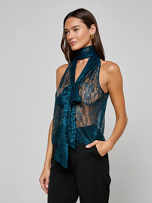A woman wears the Reece Lace Tie Neck Blouse—sleeveless, sheer teal lace with a matching tie-neck—paired with black trousers. She accessorizes with gold hoop earrings and has long brown hair, one hand casually in her pocket.