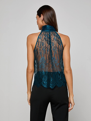 A woman with long brown hair models the Reece Lace Tie Neck Blouse, a sleeveless teal floral halter top, paired with black pants and shown from the back against a plain light gray background.