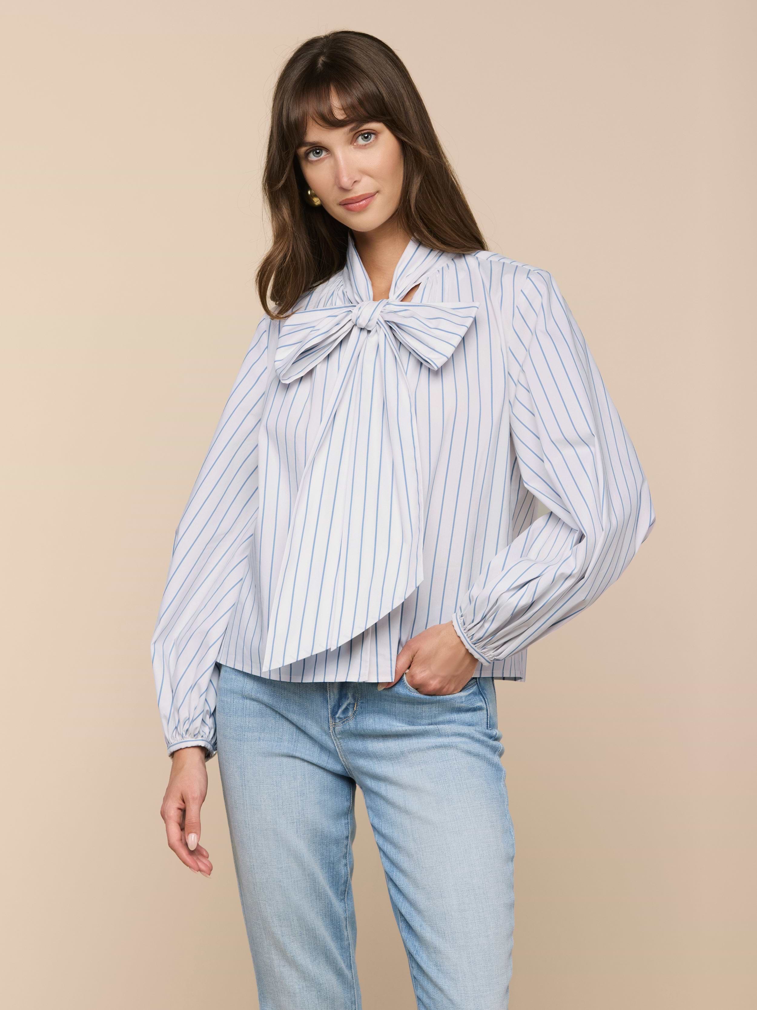 A woman models the Deon Tie-Neck Blouse, featuring white poplin with blue vertical stripes, blouson sleeves, and an oversized neck tie, paired with light blue jeans against a beige background.