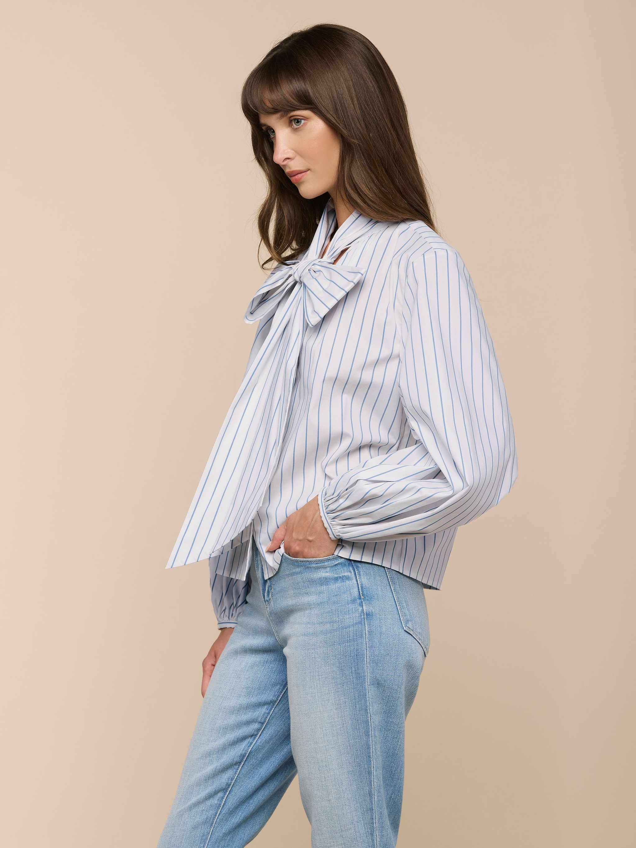 A woman with long brown hair stands in profile against a beige background, wearing the Deon Tie-Neck Blouse—an organic cotton-blend poplin top with thin blue stripes, oversized neck tie, blouson sleeves—and light blue jeans. She appears thoughtful.