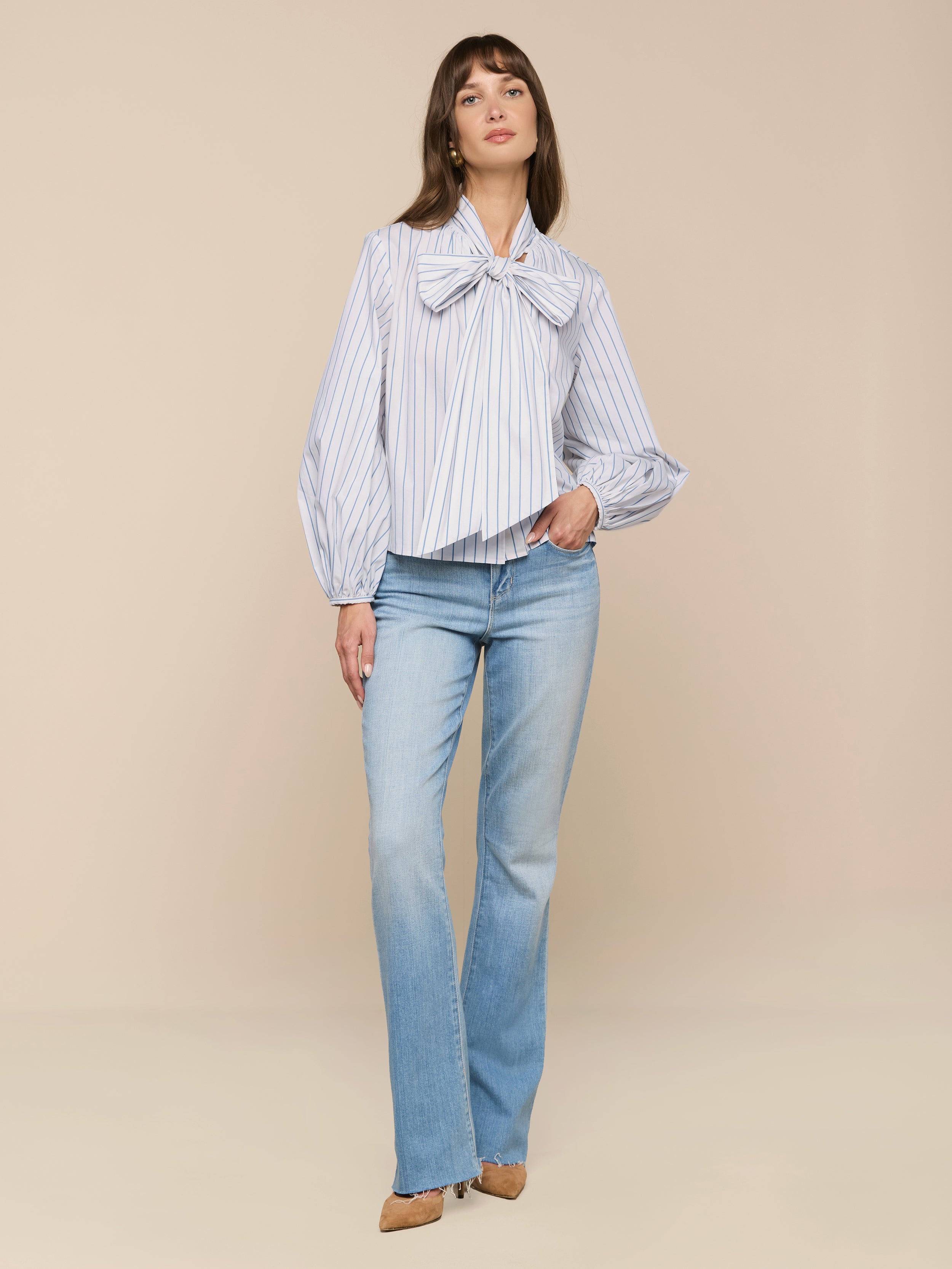 A woman stands against a beige background wearing the Deon Tie-Neck Blouse with blouson sleeves and an oversized neck tie, paired with light blue flared jeans, beige pointed shoes, and her long brown hair down, one hand in her pocket.