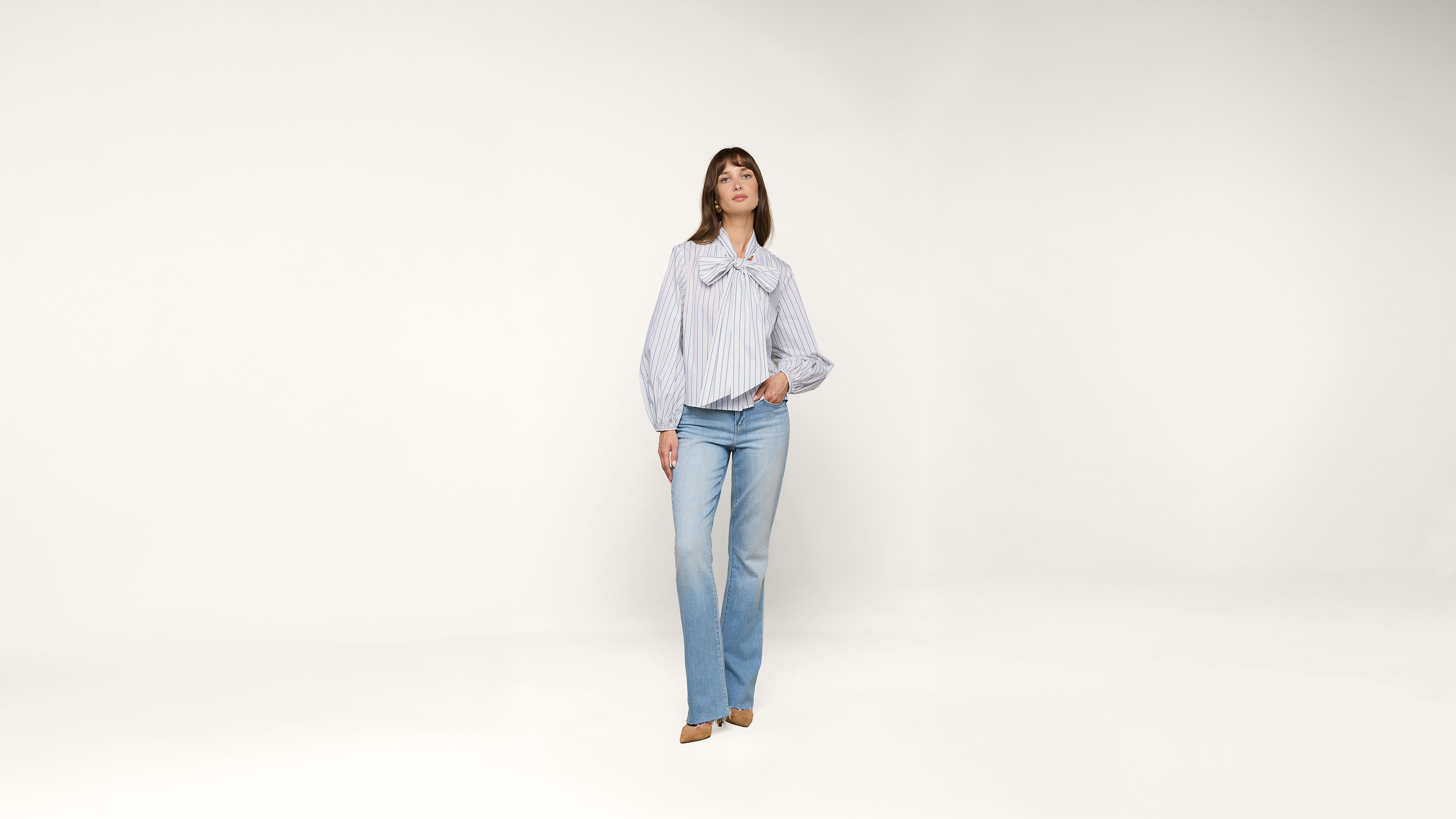 A woman stands against a plain white background, wearing a light blue and white striped blouse with a large bow and light blue jeans. She has one hand in her pocket and is looking forward.