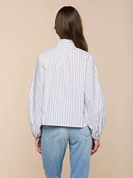 A person with long brown hair is seen from behind, wearing the Deon Tie-Neck Blouse with thin vertical stripes and blouson sleeves, paired with light blue jeans, set against a beige background.