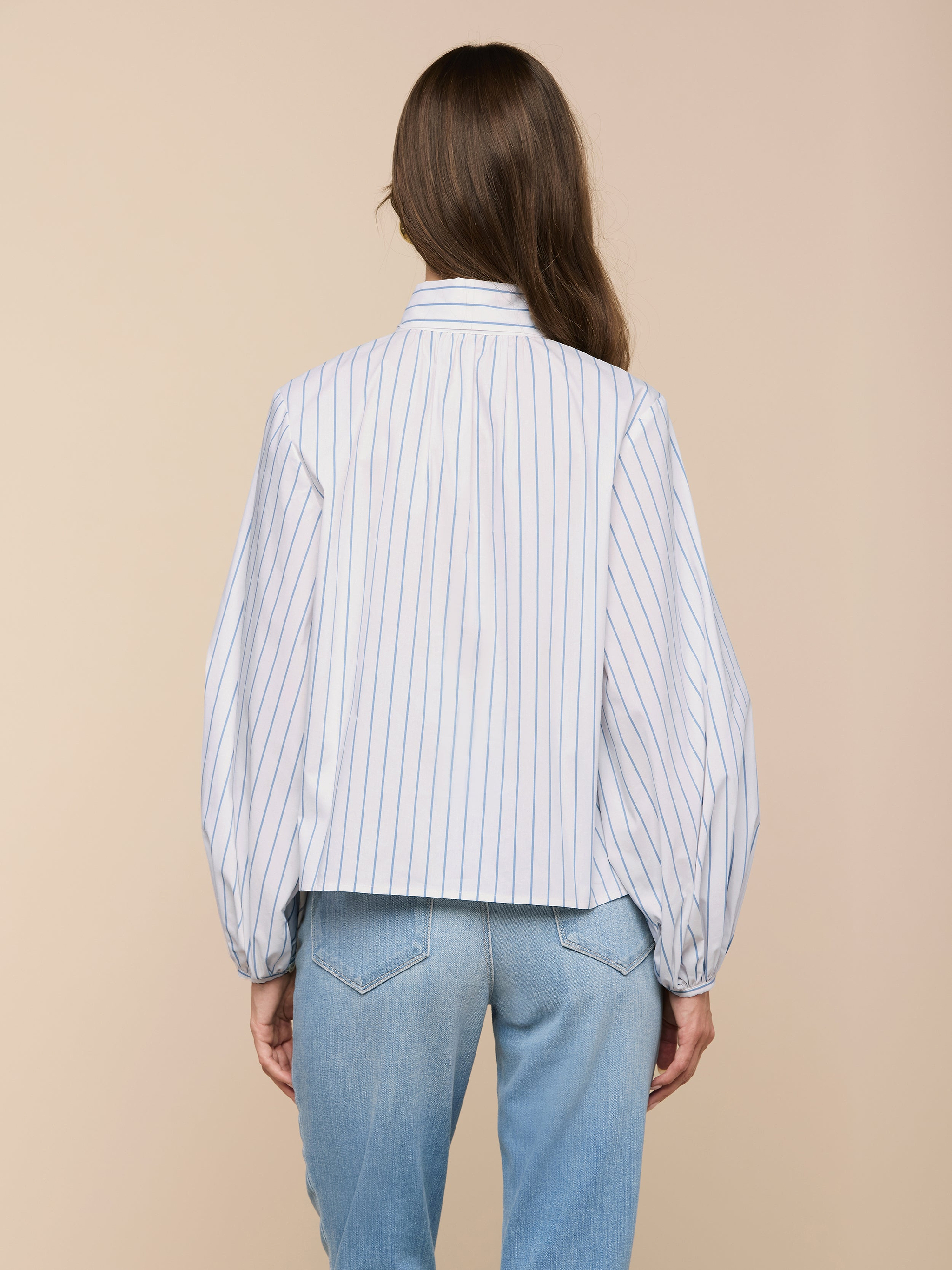 A person with long brown hair is seen from behind, wearing the Deon Tie-Neck Blouse with thin vertical stripes and blouson sleeves, paired with light blue jeans, set against a beige background.