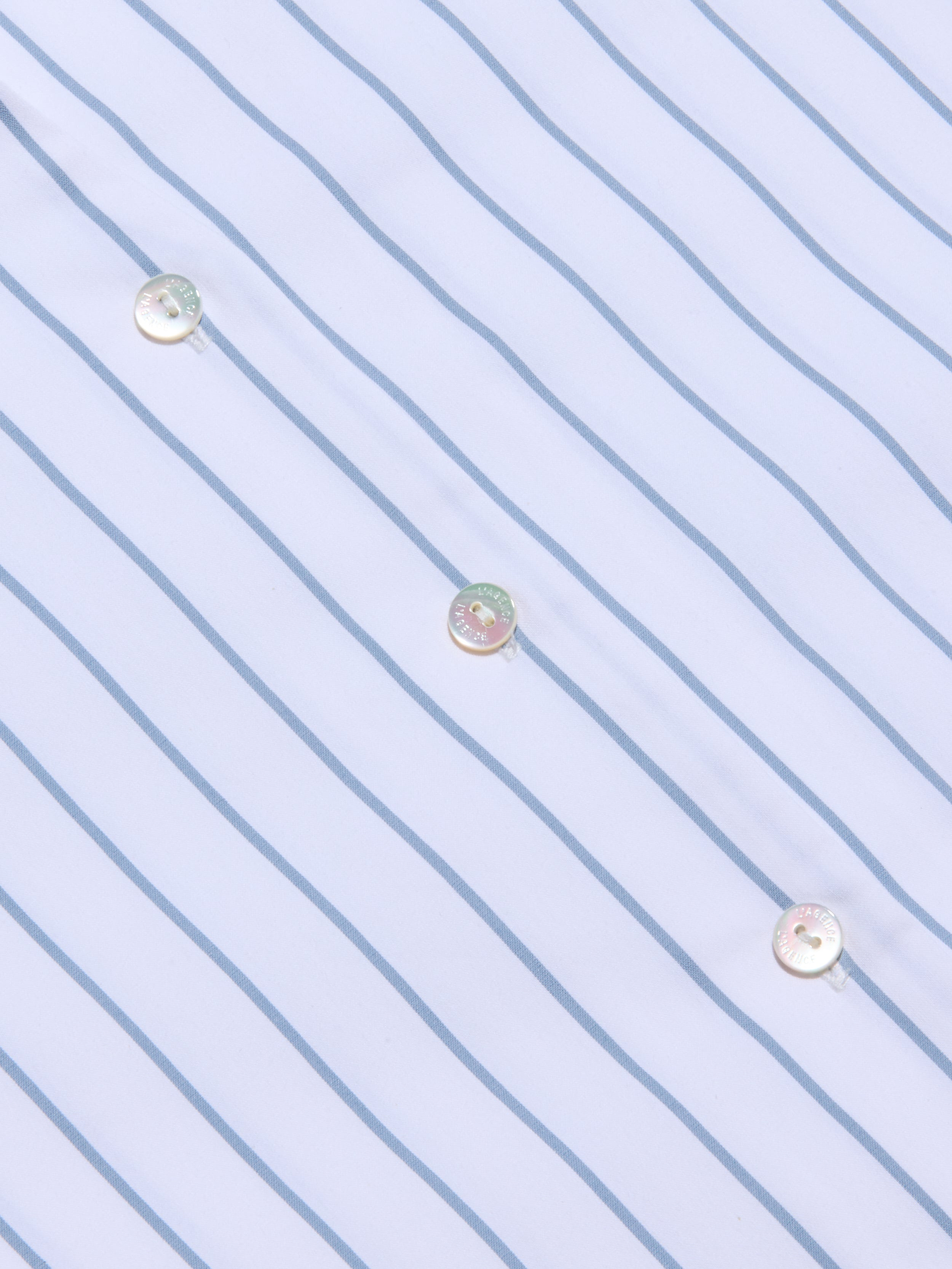 Close-up of the Deon Tie-Neck Blouse made from organic cotton-blend poplin featuring thin blue vertical stripes and three iridescent buttons sewn diagonally across the fabric.