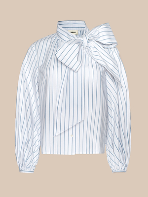 The Deon Tie-Neck Blouse is a white organic cotton-blend poplin top featuring vertical blue pinstripes, blouson sleeves, and an oversized neck tie on the right shoulder, shown against a beige background.