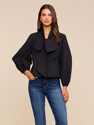 A woman with long brown hair stands against a beige background, wearing the Deon Tie-Neck Blouse—crafted from soft organic cotton-blend fabric. The black blouse features an oversized neck tie and blouson sleeves, and is paired with blue jeans. She looks at the camera with a neutral expression.