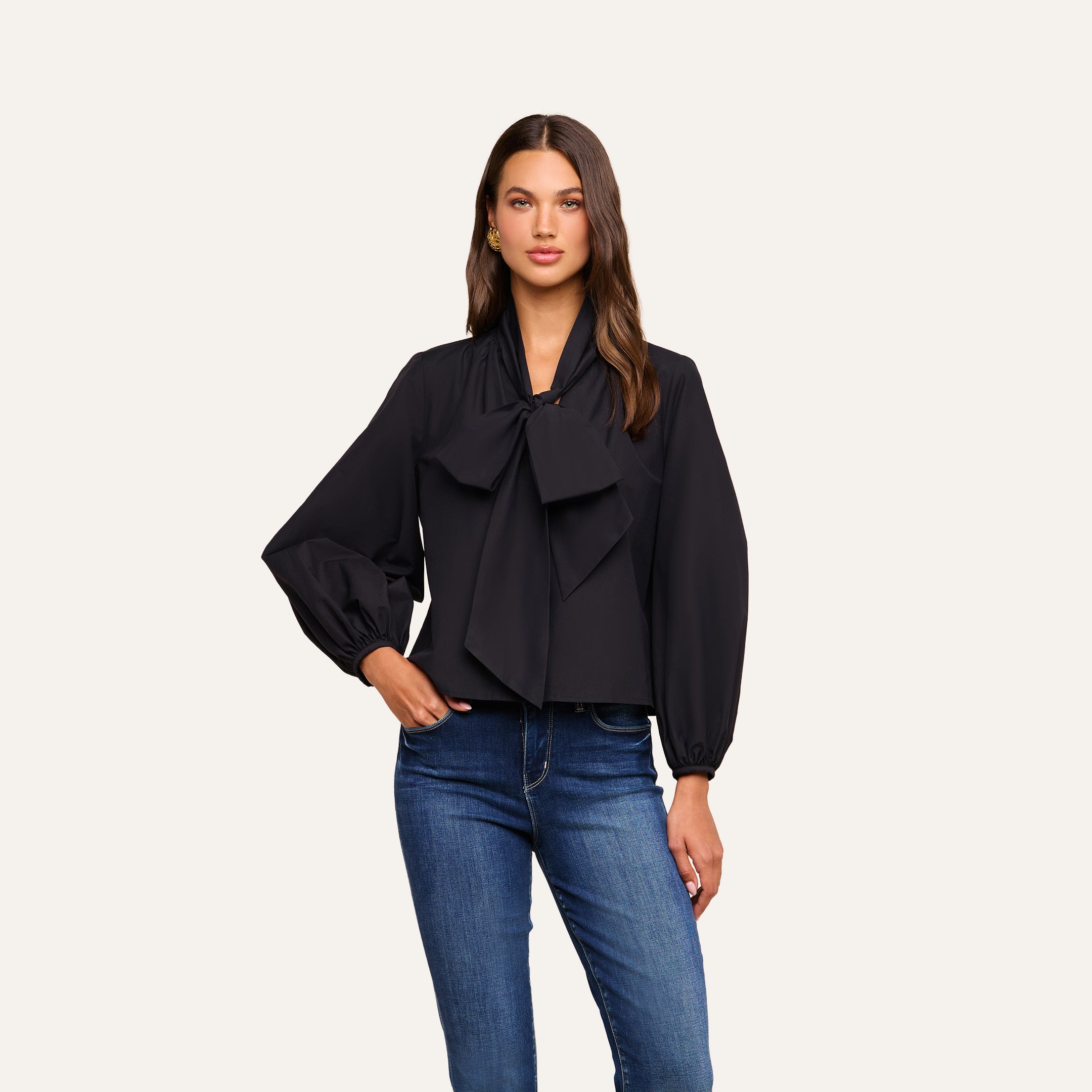 A woman is wearing the Deon Tie-Neck Blouse in black, featuring blouson sleeves and an oversized neck tie, styled with blue jeans and standing against a plain light background.