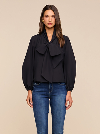 A woman stands facing forward against a plain beige background, wearing the Deon Tie-Neck Blouse—a black long-sleeve top with blouson sleeves and an oversized neck tie—paired with blue jeans.