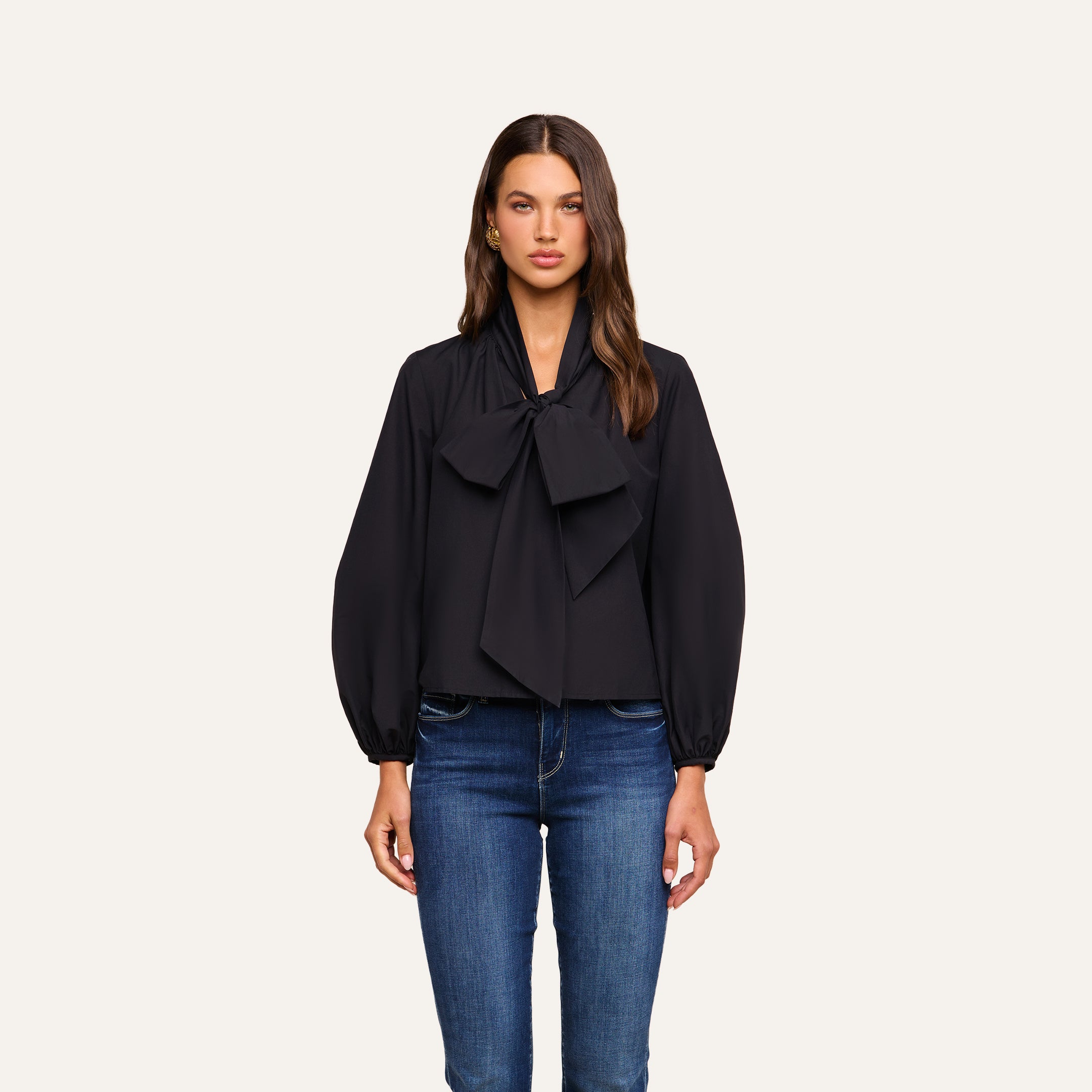 A woman wearing the Deon Tie-Neck Blouse, crafted from organic cotton poplin with oversized proportions and long blouson sleeves, paired with blue jeans and standing against a plain, light background.