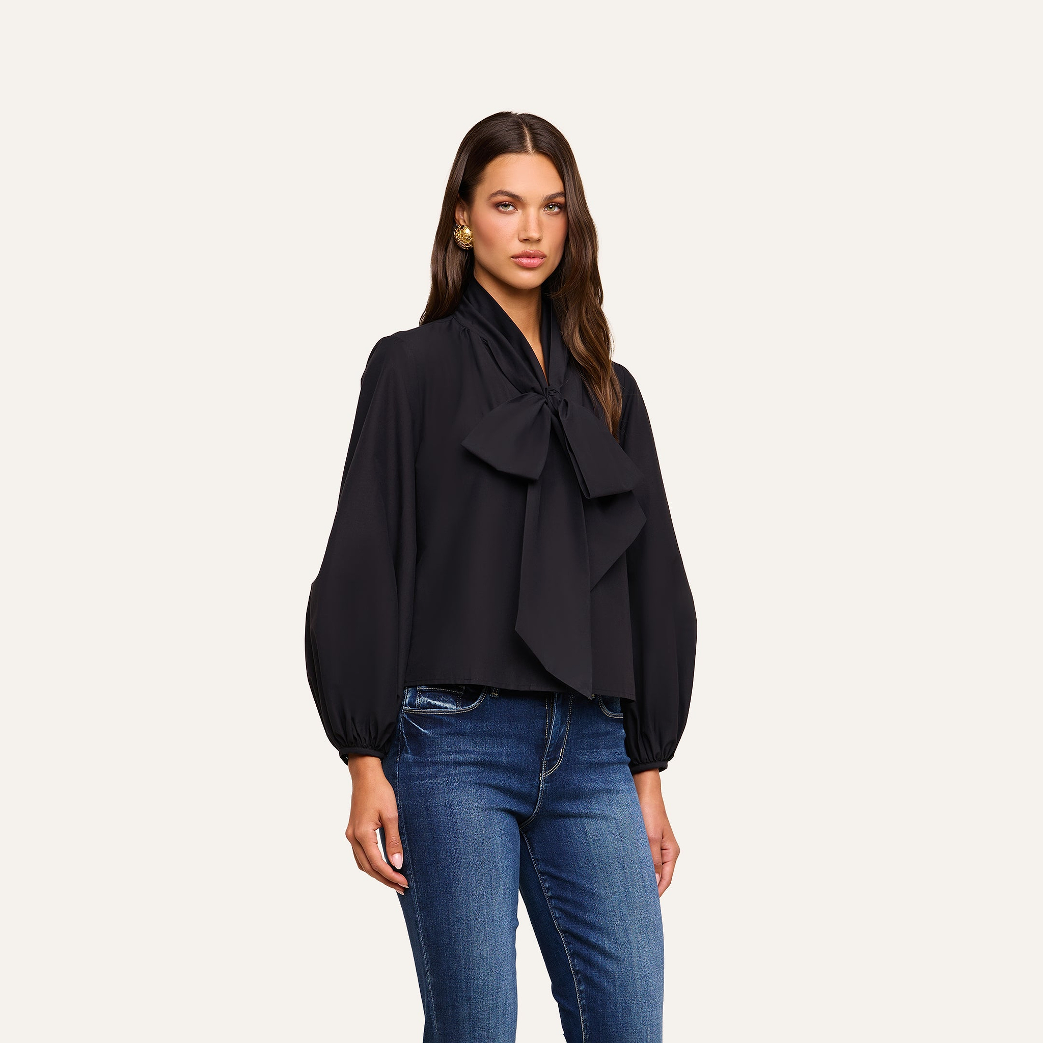 A woman wears the Deon Tie-Neck Blouse, crafted from organic cotton poplin with blouson sleeves and an oversized neck tie, paired with blue jeans and standing against a plain light background.