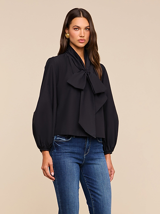 A woman wearing the Deon Tie-Neck Blouse—featuring blouson sleeves, a dramatic oversized neck tie, and crafted from an organic cotton-blend fabric in black—is paired with blue jeans and stands against a plain beige background.