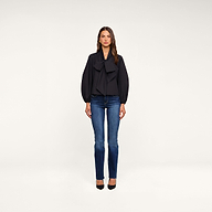 A woman stands facing forward against a plain white background, wearing the Deon Tie-Neck Blouse—an oversized blouse crafted from organic cotton poplin with blouson sleeves—paired with blue jeans and black heels.