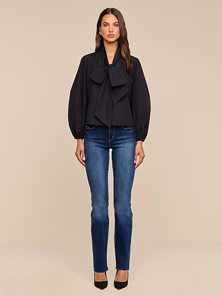 A woman faces forward against a plain beige background, wearing the Deon Tie-Neck Blouse in black with blouson sleeves and an oversized neck tie, paired with blue jeans and black heels.