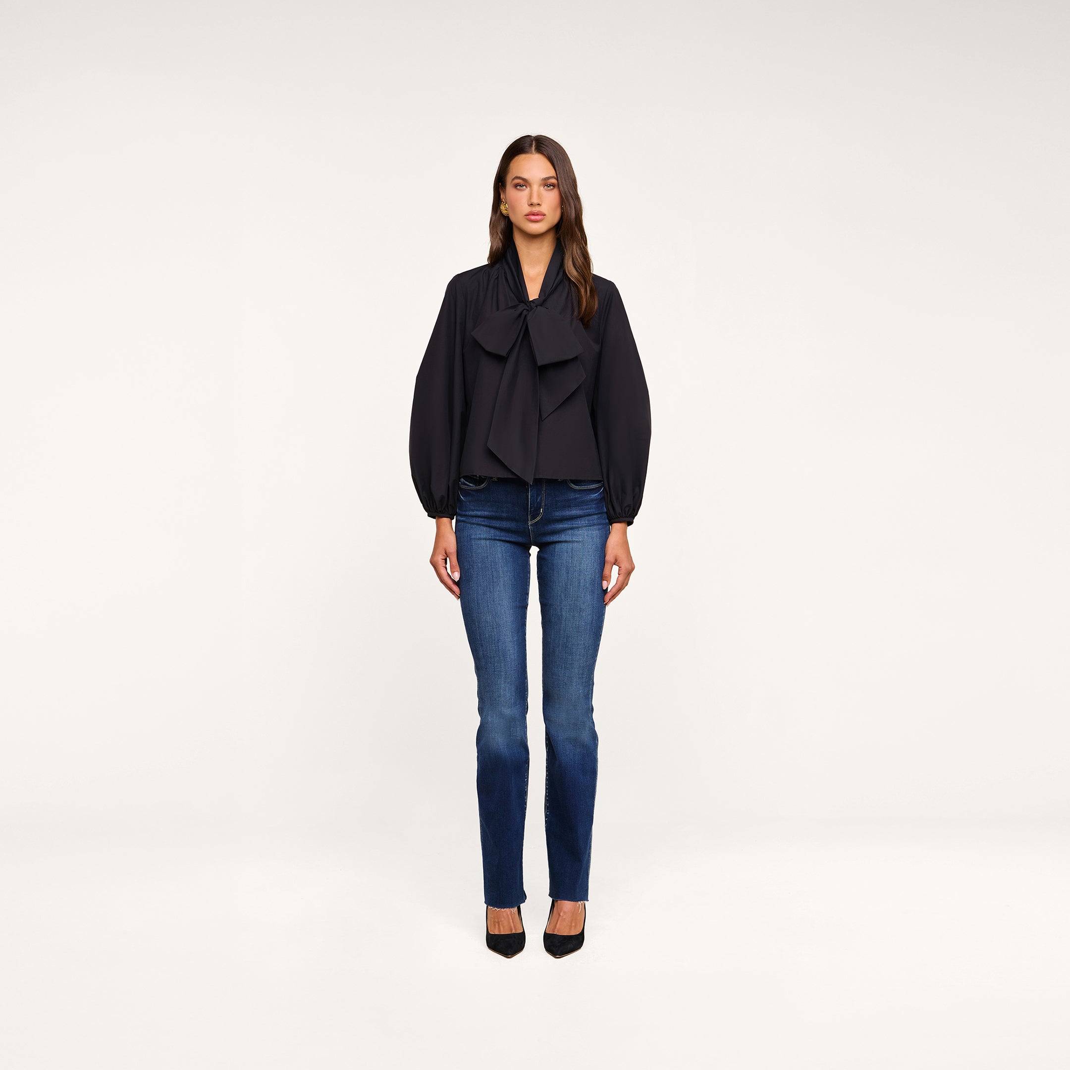 A woman stands facing forward against a plain white background, wearing the Deon Tie-Neck Blouse—an oversized blouse crafted from organic cotton poplin with blouson sleeves—paired with blue jeans and black heels.