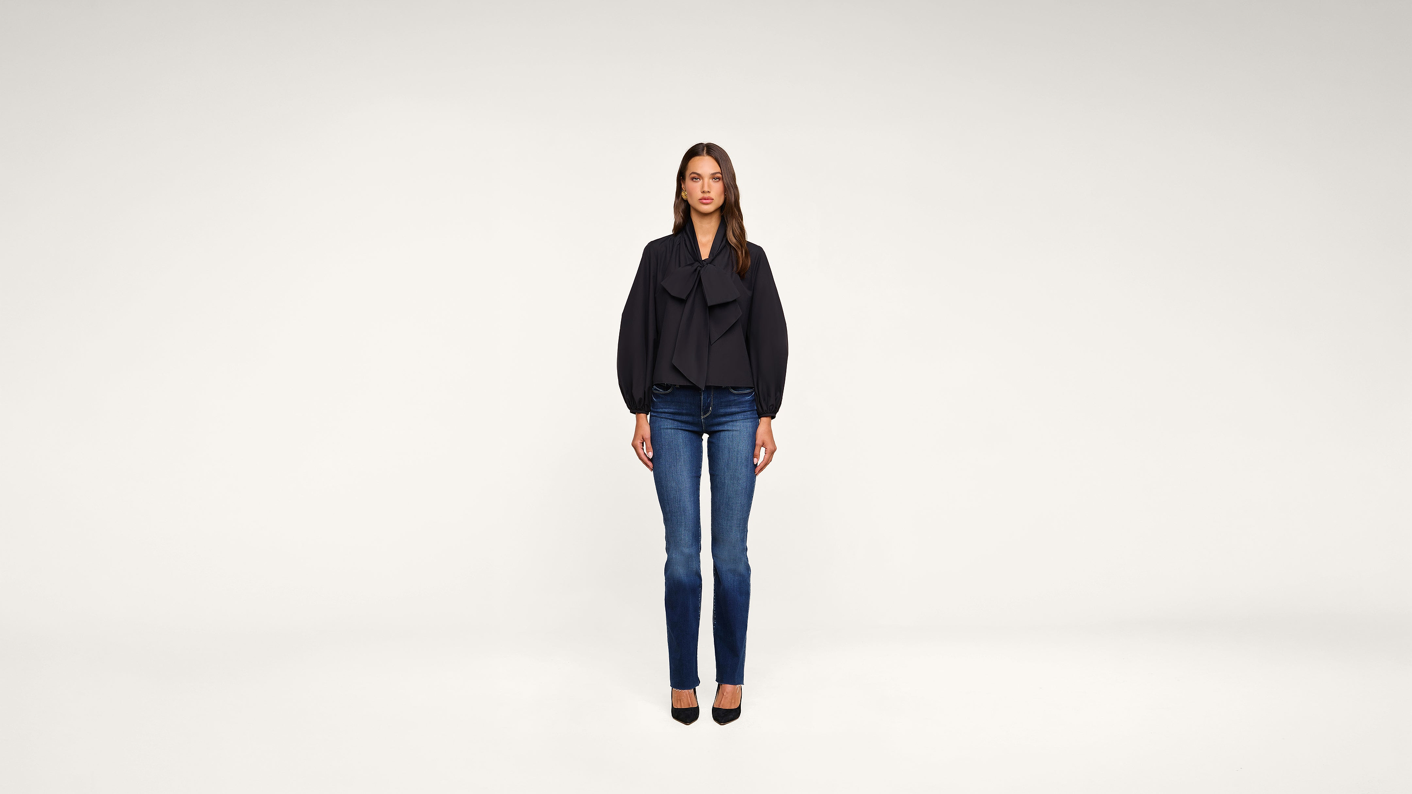 A woman stands facing forward against a plain white background, wearing a dark blouse, blue jeans, and black heels.