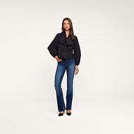 A woman stands in a studio wearing the Deon Tie-Neck Blouse—an oversized black blouse with blouson sleeves—paired with blue high-waisted jeans and black heels. She is set against a plain white background.