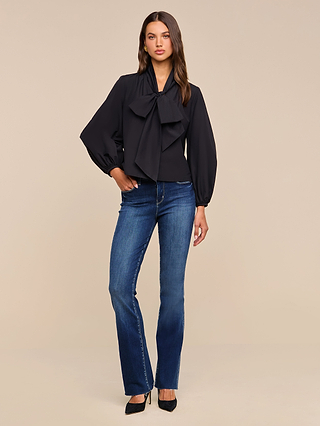 A woman stands against a beige background wearing the Deon Tie-Neck Blouse, featuring an oversized neck tie and blouson sleeves. She pairs it with blue jeans and black heels, her long brown hair styled loosely as she poses with one hand in her pocket.