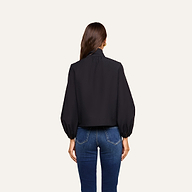 A woman with long brown hair is standing with her back to the camera, wearing the Deon Tie-Neck Blouse paired with blue jeans against an off-white background.