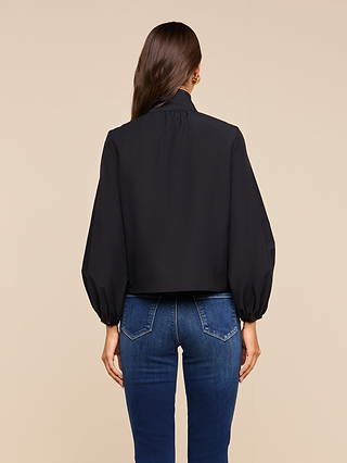A woman stands facing away, wearing the Deon Tie-Neck Blouse in black with blouson sleeves and blue jeans. The background is a solid beige color.