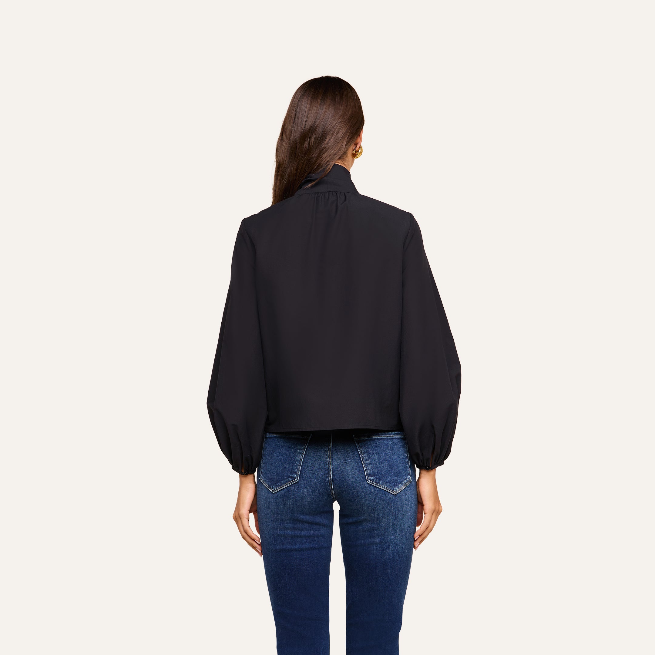 A woman with long brown hair is standing with her back to the camera, wearing the Deon Tie-Neck Blouse paired with blue jeans against an off-white background.