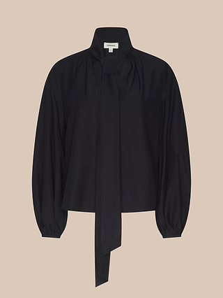 The Deon Tie-Neck Blouse, a long-sleeved black top made from an organic cotton-blend fabric, features blouson sleeves and a high neckline accented with an oversized neck tie detail, shown against a plain beige background.